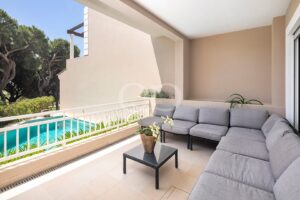 Outdoor balcony seating area with gray sectional cushions, a glass-top coffee table, and a potted plant, overlooking a pool and greenery beyond the railing.