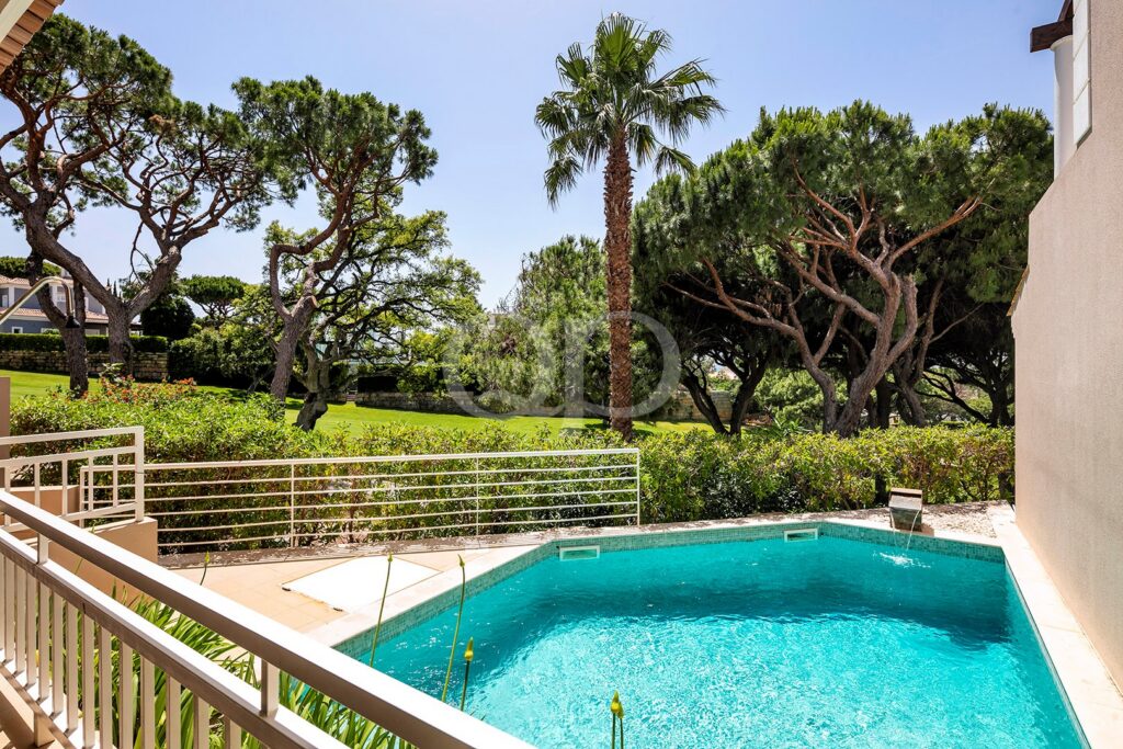 Une piscine dans un jardin, aux eaux turquoise, avec une balustrade blanche et des arbres luxuriants derrière une haie, par une journée ensoleillée.