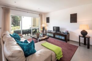 Bright living room with a beige leather sofa, turquoise cushions, and a dark wood coffee table facing a TV on a console; sliding glass doors to a balcony.
