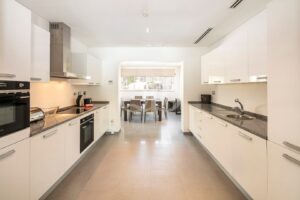 Bright, modern kitchen with white glossy cabinets, gray granite countertops, and stainless steel appliances, opening to a dining area with a table and chairs.