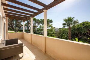 Sunlit beige balcony with wicker seating and a wooden pergola, overlooking trees and a palm in a sunny courtyard.