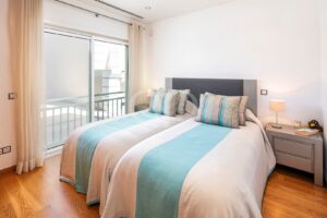 Bright bedroom with a double bed, teal-and-white duvet, striped pillows, and matching nightstands by a sliding door to a balcony.