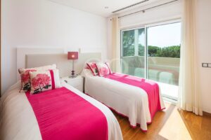 Twin bedroom with white and hot-pink bedding, pink floral pillows, and a glass door to a sunny balcony.