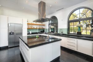 Modern white kitchen with a central island and stainless steel range hood, large arched windows, and black countertops/Sleek appliances.