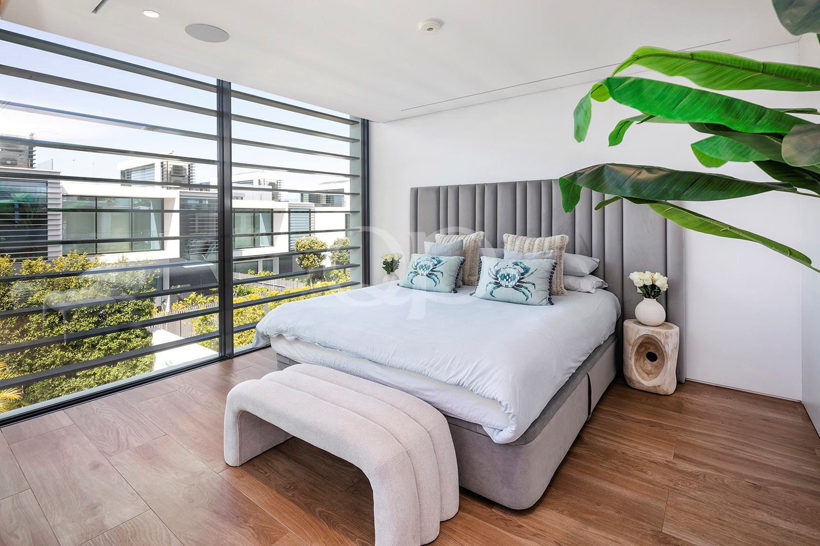 Helles, modernes Schlafzimmer mit einer großen Glasfassade, einem grauen Polsterbett und einer tropischen Pflanze auf der rechten Seite.