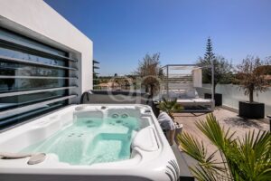 Ein weißer Whirlpool im Freien auf einer Holzterrasse mit einer modernen weißen Wand, einer Lounge-Cabana und Topfpflanzen unter strahlend blauem Himmel.