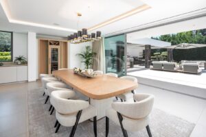Ein offener Speisesaal mit einem langen Holztisch, beigen Stühlen und einem modernen Kronleuchter, der sich zu einer sonnendurchfluteten Terrasse im Freien hin öffnet.