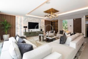 Ein luxuriöses Wohnzimmer mit einer weißen Eckcouch, grauen Kissen und einer Marmorwand hinter einem Flachbildfernseher.