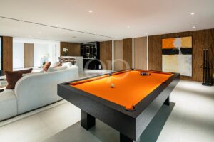 Ein luxuriöser, moderner Keller mit einem orangefarbenen Billardtisch im Vordergrund und einem weißen, geschwungenen Sofa im Hintergrund.