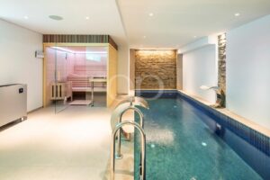 Hallenbadbereich mit einer Sauna mit Glasfront auf der linken Seite, einer Akzentwand aus Stein am anderen Ende und sanfter Deckenbeleuchtung für eine Wellness-Atmosphäre