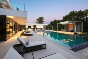 Ein luxuriöser, moderner Garten mit einem langen, rechteckigen Pool, weißen Liegestühlen und einem Haus mit Glasfassade in der Abenddämmerung.