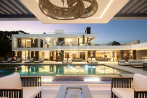 Ein luxuriöses, zweistöckiges, modernes Haus mit einem rechteckigen Pool, weißen Terrassenmöbeln und warmer Innenbeleuchtung in der Abenddämmerung.