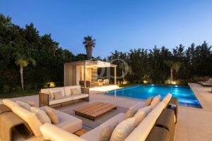 Eine luxuriöse Terrasse am Pool in der Abenddämmerung mit cremefarbenen Sofas, einem Couchtisch aus Holz und einer modernen Cabana inmitten von Palmen und Hecken.