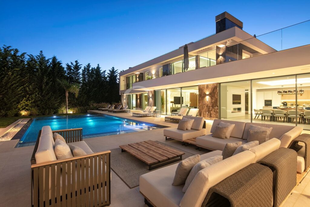 Moderne zweistöckige Villa in der Abenddämmerung mit beleuchteten Innenräumen, einem rechteckigen Pool und Sitzgelegenheiten rund um die Terrasse.
