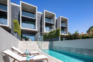 Contemporary apartment complex with glass balconies overlooking a rectangular pool and blue sky.
