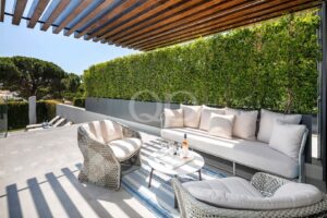 Modern outdoor lounge area with cream cushions, wicker chairs, a marble table, a bottle of wine and glasses under a slatted wooden pergola next to a green hedge.