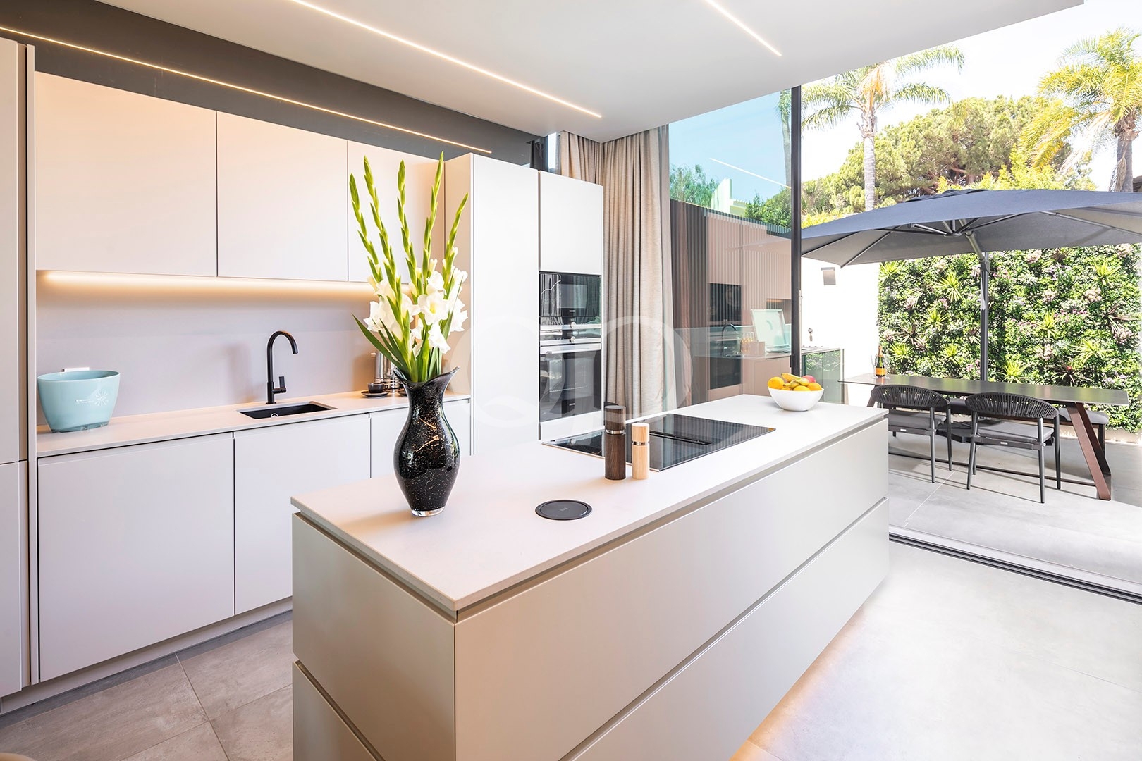 Bright, modern white kitchen with a large island, built-in oven, and minimalist cabinets; outdoor dining area visible through glass doors.