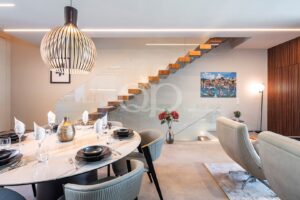 Round glass dining table set with black bowls, crystal glasses, and folded napkins in a bright modern living-dining space with a sculptural pendant light and glass-railed staircase