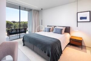 Modern bedroom with a large glass balcony door, view of trees, and a gray bed with blue-gray throw and pillows