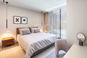 Modern bedroom with a large bed, gray upholstered headboard, beige bedding, two abstract framed prints, and vertical blinds by a floor-to-ceiling window.