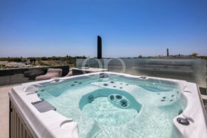 Outdoor rooftop hot tub on a sunlit patio with glass railing and lounge seating, under a clear blue sky.