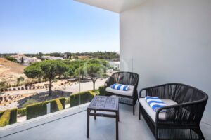 Balcony seating area with two black wicker chairs, a small table, and blue striped towels, overlooking a sunlit landscape and trees.