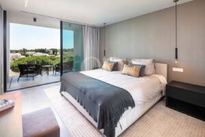 Hotel room with a large bed, gray throw, and decorative pillows; sliding glass door opens to a balcony with chairs and a table overlooking greenery and houses far away.