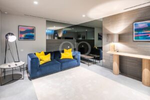 Modern lounge with a blue sofa and bright yellow cushions, a tripod floor lamp, and a marble-topped side table near an abstract colorful painting on the wall.