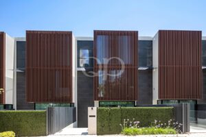 Modern townhouse facade with brown vertical wooden slats covering large windows, white and dark gray walls, and a trimmed hedge against a blue sky.