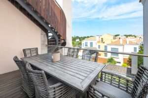 Balcon extérieur avec une table en bois gris et des chaises en osier, donnant sur un quartier résidentiel.