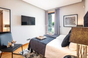 Deux lits d'hôtel séparés, recouverts de couettes matelassées grises et flanqués de serviettes pliées, face à une télévision fixée au mur, dans une chambre lumineuse et moderne.