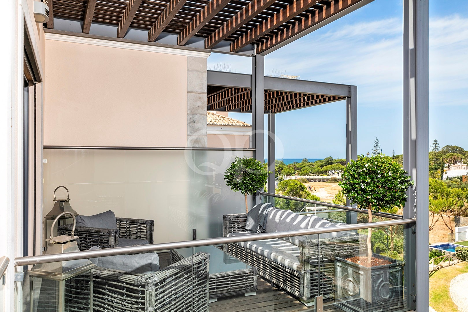Terrasse extérieure avec des chaises en osier gris, une balustrade en verre et des arbres en pot, donnant sur la côte et la verdure.