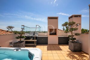 Une terrasse sur le toit avec un jacuzzi, des chaises longues en osier et des arbres en pot, le tout sur fond de ciel bleu.