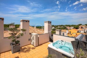 Une terrasse sur le toit avec un jacuzzi, un sol en terre cuite et un arbre en pot, donnant sur une ville méditerranéenne et la mer bleue sous un ciel dégagé.
