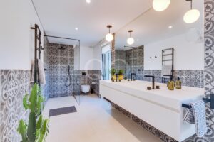 Modernes Badezimmer mit einem langen, weißen Doppelwaschtisch, schwarzen Armaturen und gemusterten grauen Fliesen an den Wänden und im Duschbereich