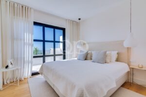 Ein helles, luftiges Schlafzimmer mit einer großen, schwarz gerahmten Glastür bzw. einem Fenster mit Meerblick, weißen, durchsichtigen Vorhängen und einem weißen, gesteppten Bett mit grauen und beigen Zierkissen. An der Decke hängt neben einem kleinen Nachttisch eine weiße Pendelleuchte.