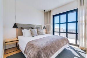Modernes Schlafzimmer mit einem großen, blau gerahmten Fenster, neutraler Bettwäsche und einer grauen Überwurfdecke in einem sonnendurchfluteten Raum