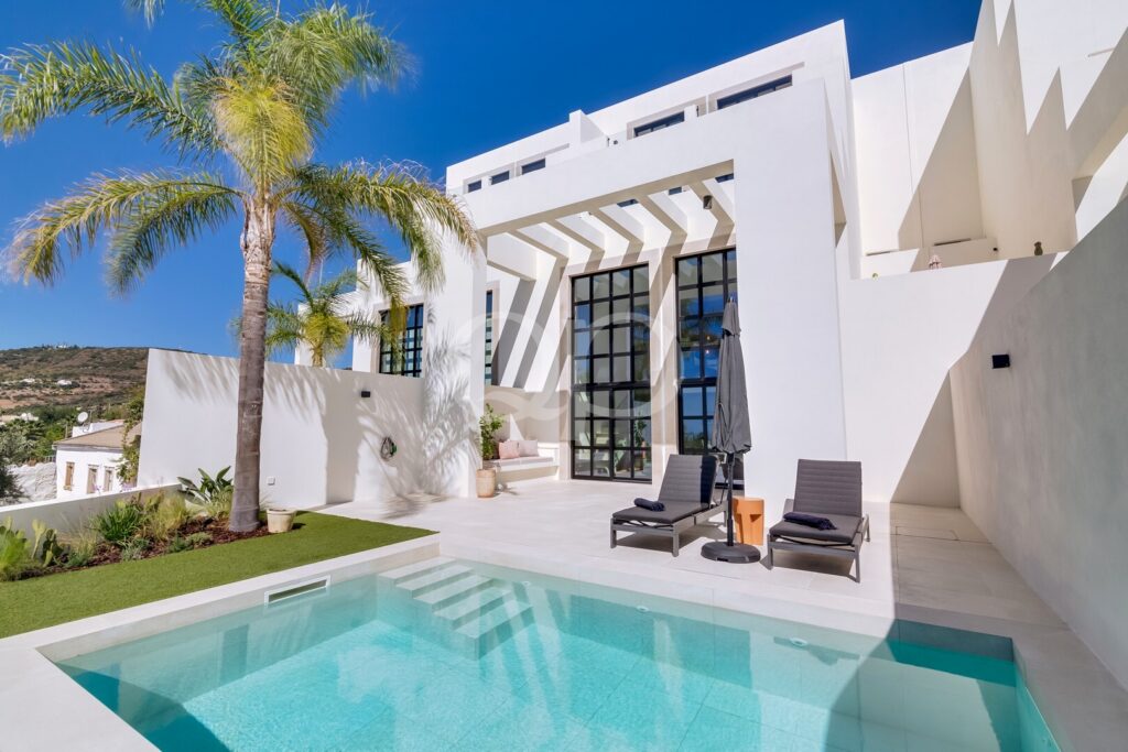 Moderne weiße Villa mit einem rechteckigen Pool, einer Palme und zwei Liegestühlen auf einer sonnigen Terrasse.