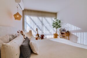 Bright bedroom with white bedding, a wicker pendant lamp, sheer curtains, and a small desk near a large window. Guitar and plants sit by the window.