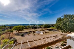 Sunny desert garden with a circular white pool, dry lawn, shrubs, and distant hills under a bright blue sky; wooden railing in the foreground.