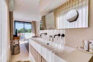 Bright modern bathroom with double sinks, black wall-mounted faucets, large mirror, and a sliding glass door to a balcony.