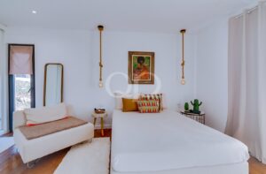 Bright minimalist bedroom with a white bed, colorful pillows, and a painting above; rope pendants flank the bed for a playful touch.