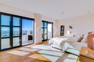 Helles, modernes Schlafzimmer mit großen Glasschiebetüren, die auf einen Balkon führen und den Blick auf den blauen Himmel und das Meer dahinter freigeben, mit weißer Bettwäsche und hellem Holzboden.