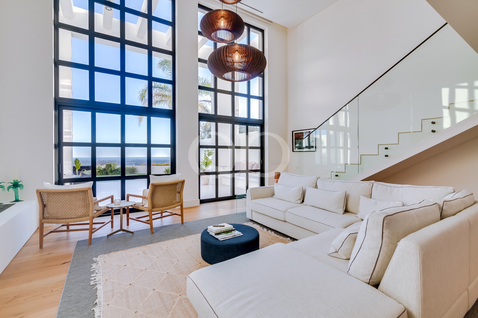Helles, modernes Wohnzimmer mit hohen, schwarz gerahmten Gitterfenstern, die den Blick auf das Meer freigeben, einem beigen Ecksofa, Korbstühlen und einer Treppe mit Glasgeländer, über der Pendelleuchten hängen.