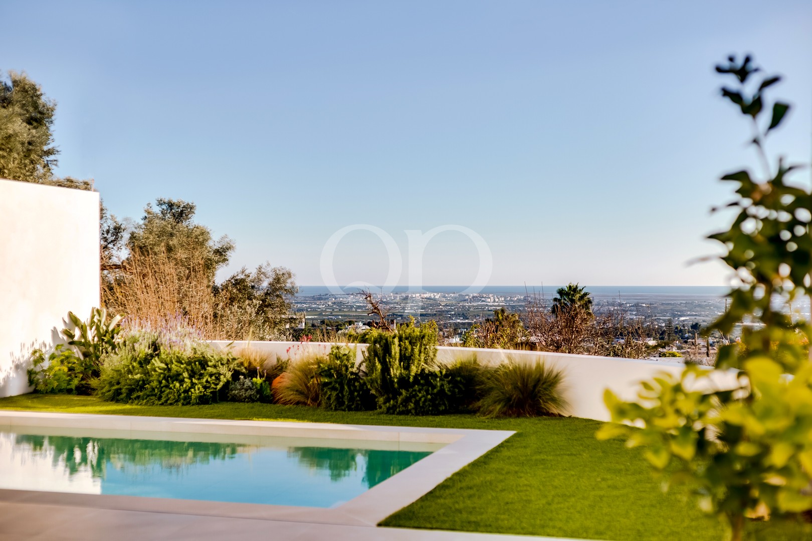 Garten am Pool mit einem rechteckigen Pool, einem gepflegten Rasen und üppiger Bepflanzung, mit Blick auf eine in der Ferne gelegene Küstenstadt und das Meer.