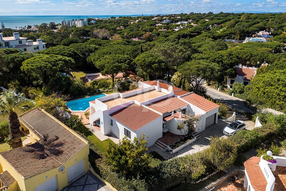 Vista aérea de uma casa de estuque branco com telhado de telhas vermelhas, piscina e jardim mediterrânico num bairro costeiro.
