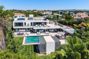 Eine moderne weiße Villa mit mehreren Terrassen, Glasgeländern und einem langen, rechteckigen Pool, der von Liegestühlen und Palmen umgeben ist.