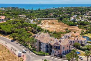 Aerial view of a coastal residential neighborhood with a large undeveloped dirt lot at center and the sea in the distance.