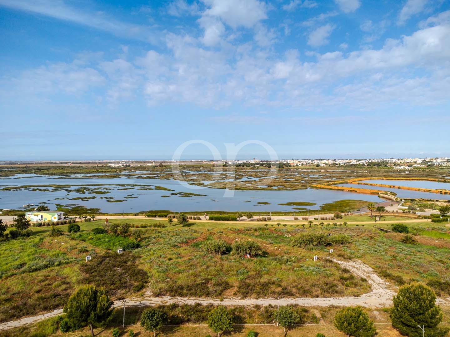 Vista ampla de zonas húmidas costeiras com poças de maré, uma cidade distante no horizonte e um céu azul brilhante acima.