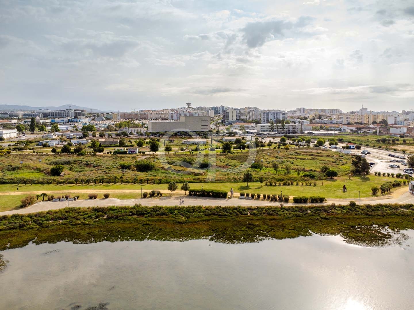 Vista aérea de um parque à beira-rio com relva verde, caminhos pedonais e uma paisagem urbana de edifícios baixos à distância, sob um céu nublado.